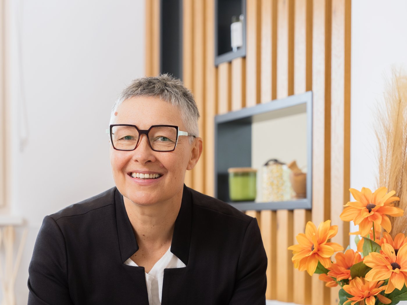 Immobilienmaklerin lacht am Besprechungstisch, im Hintergrund Holzlamellen und orange Blumen – Mitarbeiterporträt für REMAX Expert